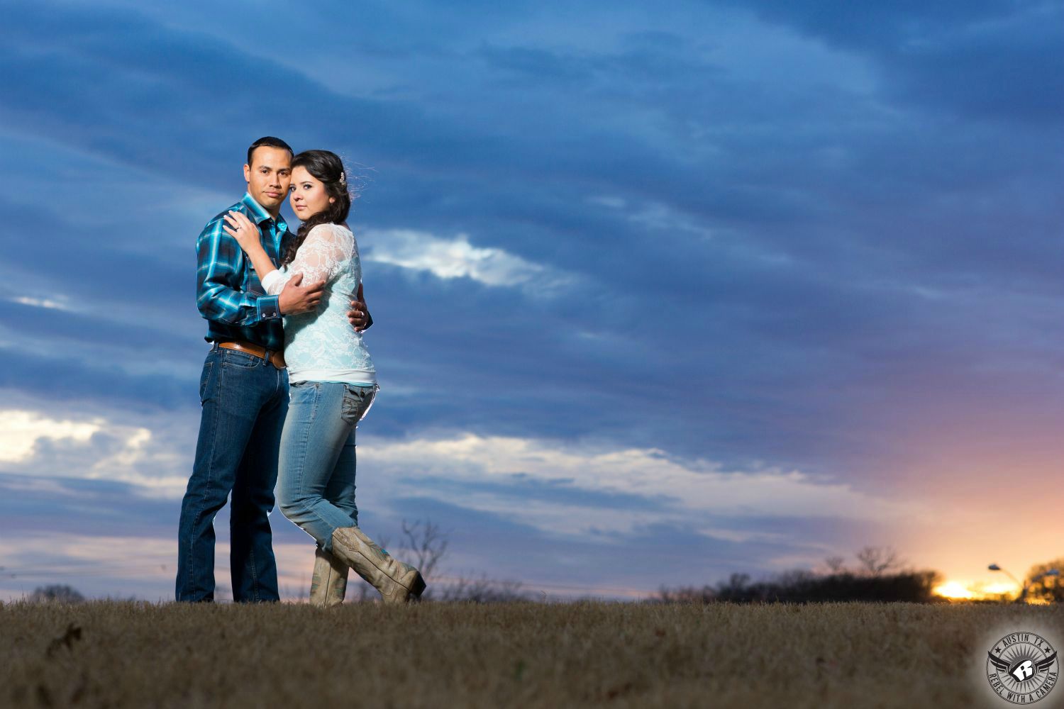 hot blond girl with blue dress and tan heals shares a cupcake with a dark haired guy wearing a dark blue shirt and black pants sitting together on the bumper of the original hey cupcake silver airstream travel trailer with a large cupcake on top in this engagement photograph in southern austin  